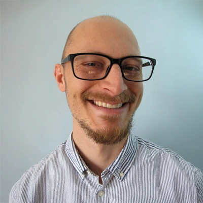 A person wearing glasses and a collared shirt is shown from the shoulders up against a light blue background