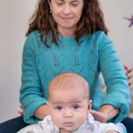 An adult wearing a teal knitted sweater sits behind an infant